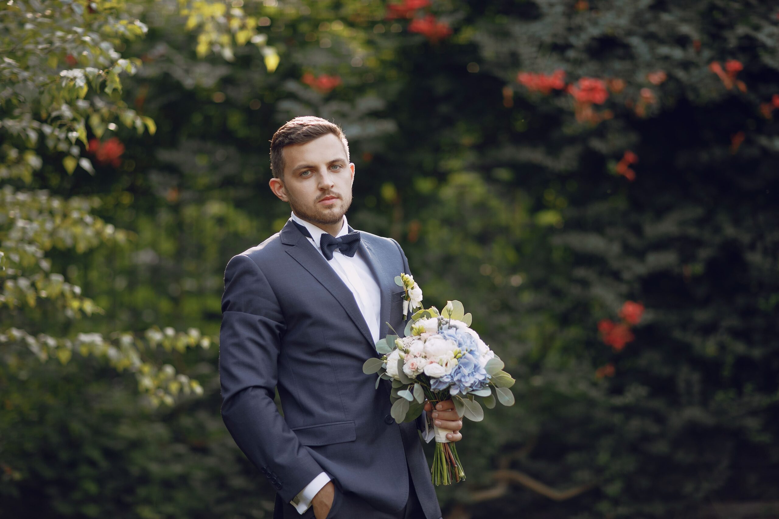handsome-stylish-groom-blue-suit-stands-park-near-bushes-with-bouquet
